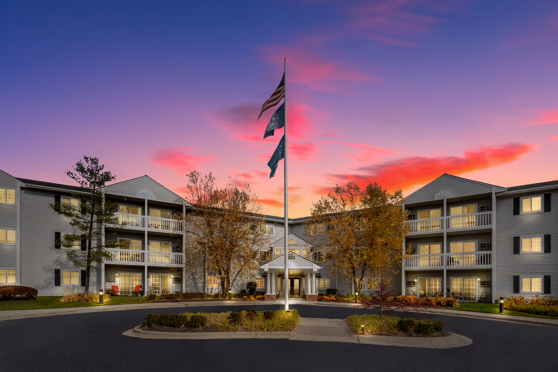 Front exterior of a three-story senior living building with balconies, a central entrance, flagpole, and landscaped circular driveway at sunset.