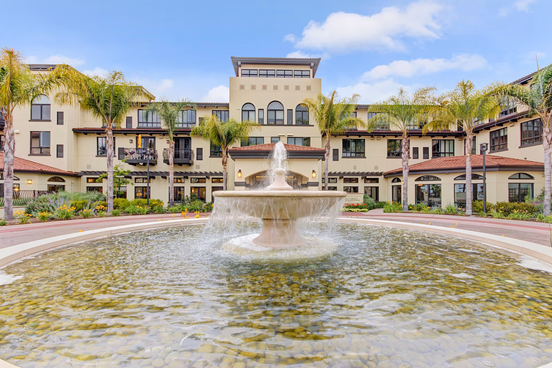Front exterior view of a multi-story senior living facility with a central water fountain surrounded by a circular pool, palm trees, and landscaped greenery under a blue sky with some clouds.
