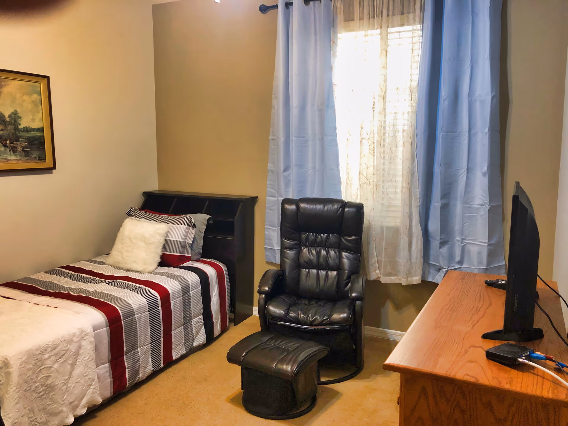 A small bedroom with a single bed covered in a striped red, white, and gray comforter with a white fluffy pillow. Next to the bed is a black leather recliner chair with a matching footrest. A wooden dresser with a flat-screen TV on top is positioned opposite the bed. The window behind the chair has sheer white curtains and light blue drapes. A framed painting hangs on the wall above the bed.