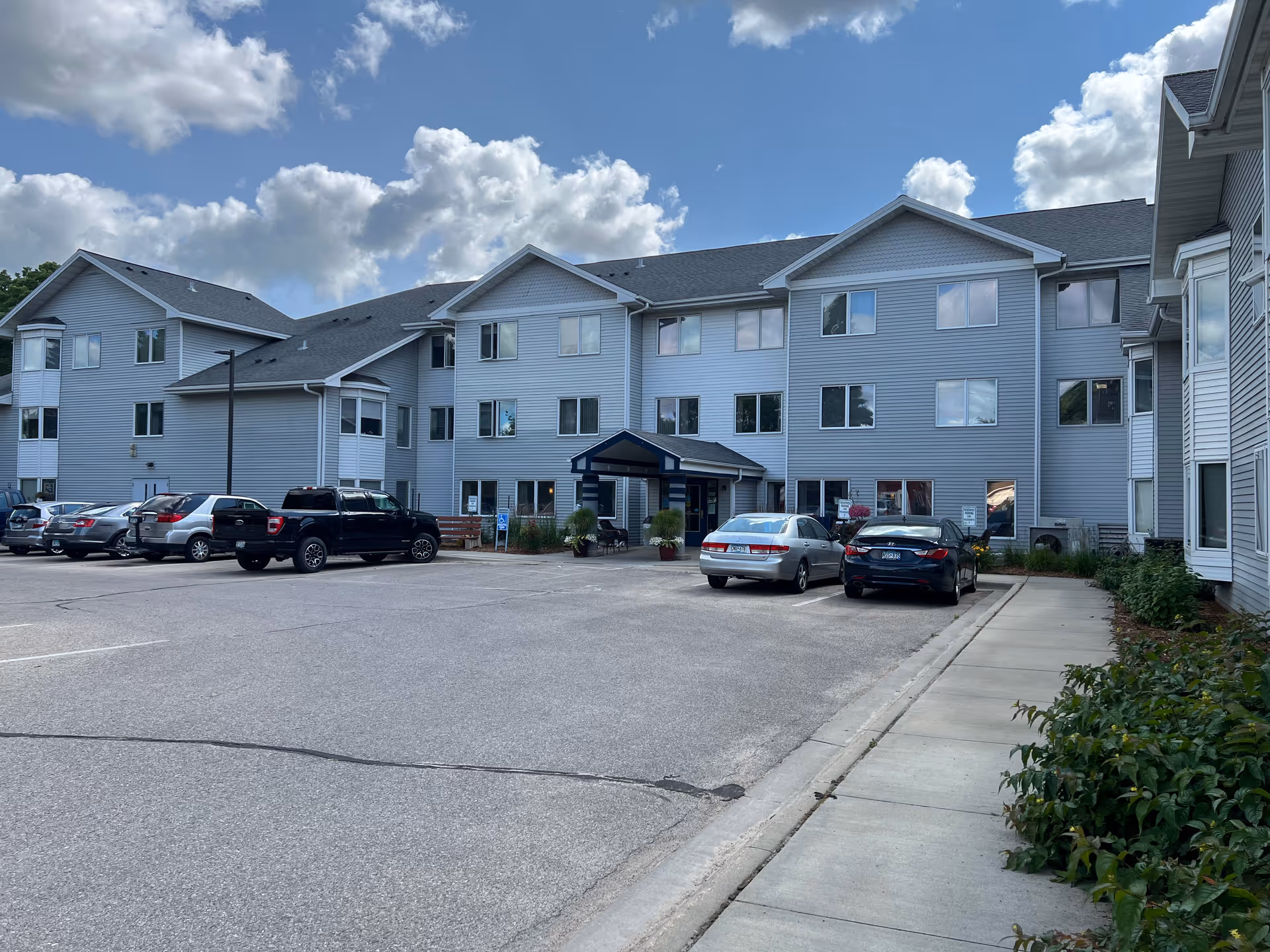 Exterior view of a three-story senior living facility building with gray siding and a dark roof under a partly cloudy sky. Several cars are parked in the parking lot in front of the building, and a sidewalk with some greenery runs along the right side.