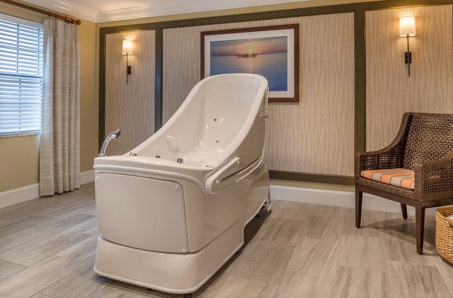 A modern walk-in bathtub sits in a softly lit room with a wicker chair, framed artwork, and a window with curtains.
