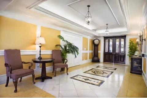 Bright furnished lobby/entryway with two upholstered chairs and a small table, decorative floor tiles, a grandfather clock, and double wooden doors.