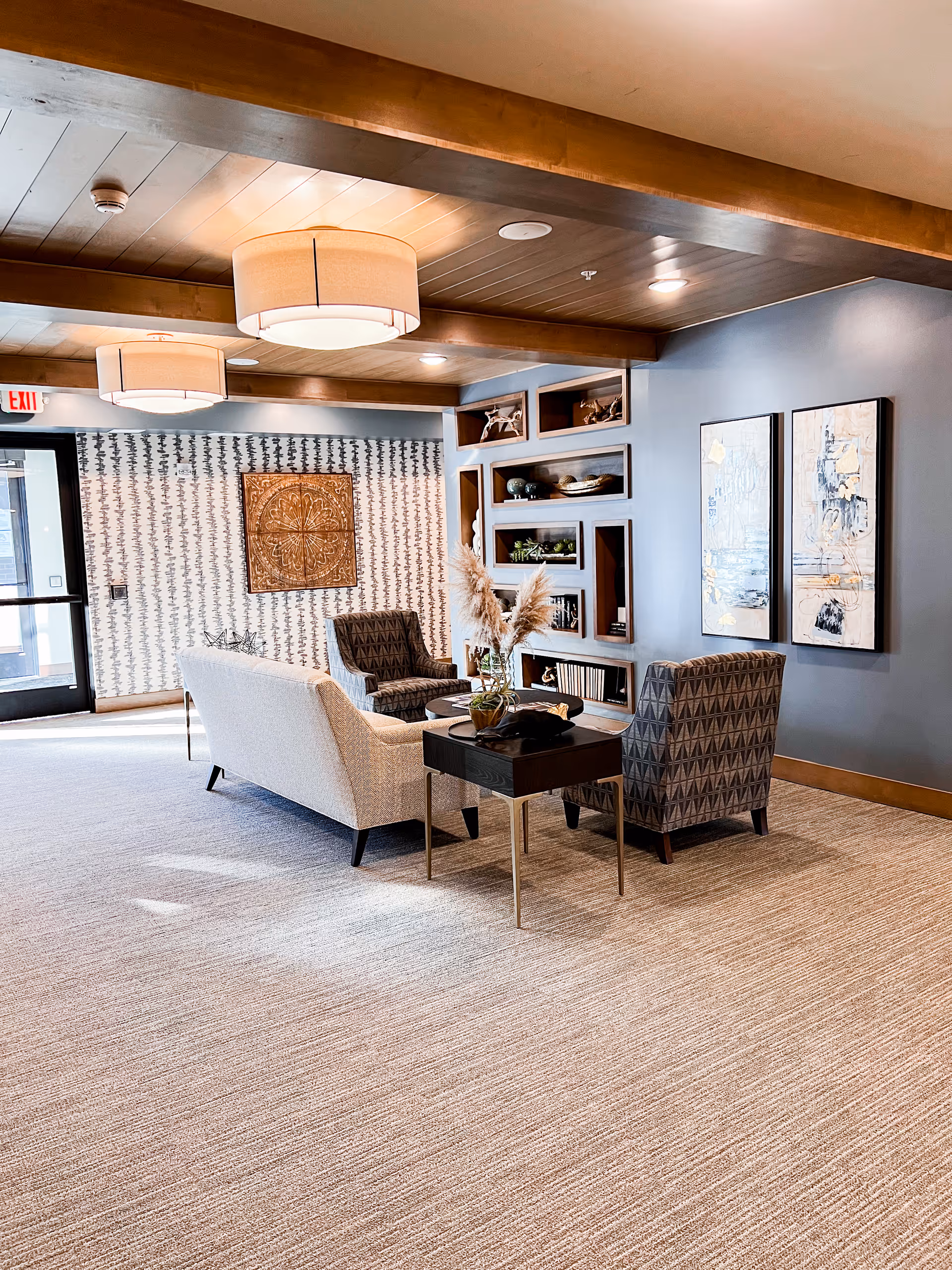 A cozy senior living facility lounge area with a beige sofa, two patterned armchairs, a small black side table with a decorative vase holding pampas grass, built-in shelves with decorative items, and two abstract paintings on a blue wall. The ceiling has wooden beams and two large round light fixtures, and the floor is carpeted.