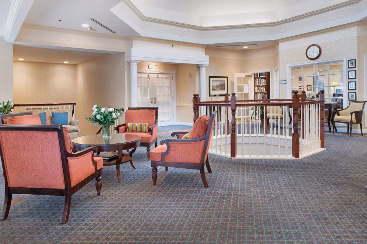A spacious and well-lit common area in a senior living facility featuring a seating arrangement with four orange upholstered chairs around a round wooden table with a vase of flowers. The room has patterned carpet flooring, cream-colored walls, and white columns framing a doorway. There is a wooden railing around a stairwell or open space in the center, a clock on the wall, framed pictures, and a glimpse into a room with bookshelves and additional seating.