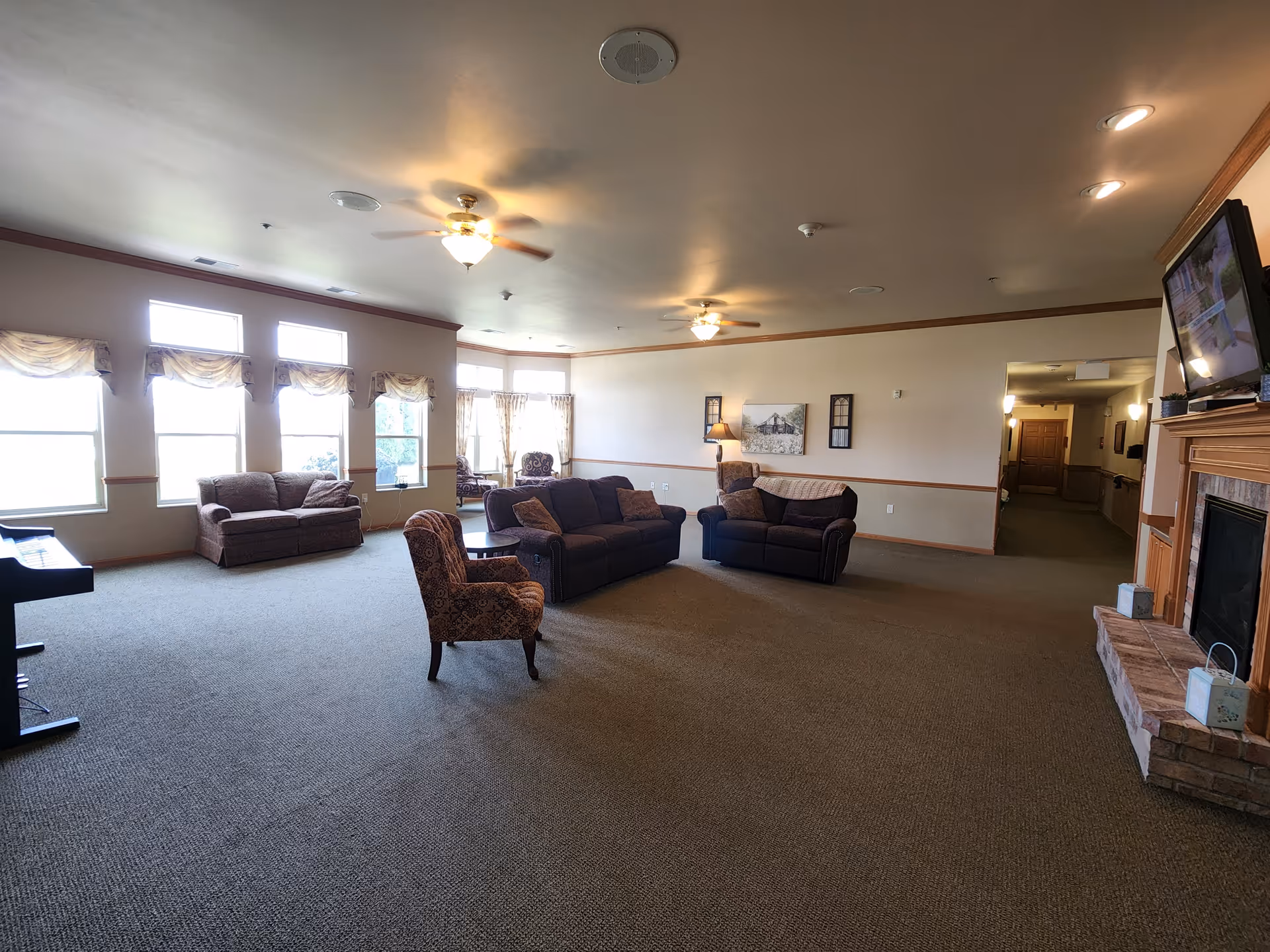 Spacious communal living room with sofas, armchairs, large windows, ceiling fans, and a fireplace.