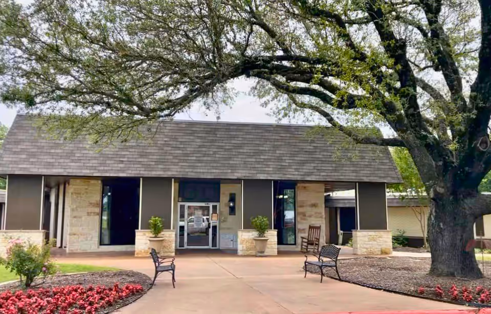 Front exterior view of North Park Health and Rehabilitation Center building with a large tree on the right, two benches on the walkway, flower beds, and a covered entrance with glass doors.