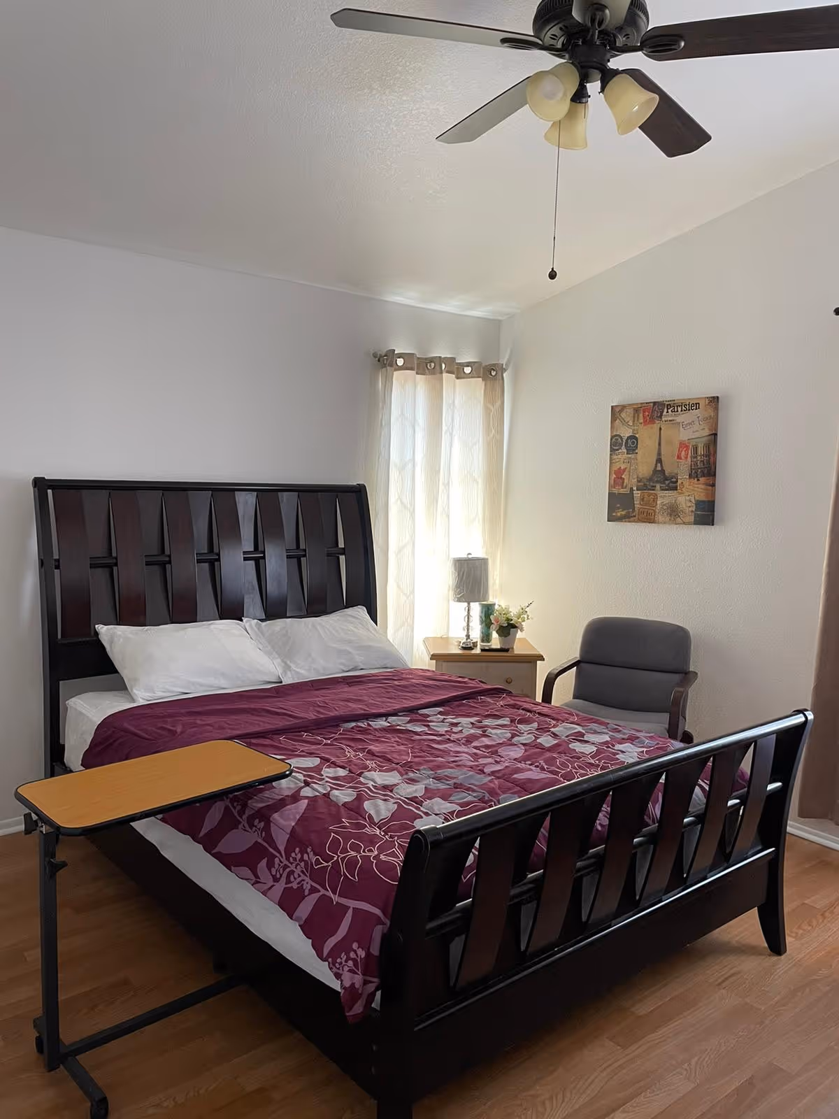 A furnished bedroom with a dark wood bed dressed in a purple floral comforter, nightstand and lamp by a curtained window, a chair, ceiling fan, and an overbed table.