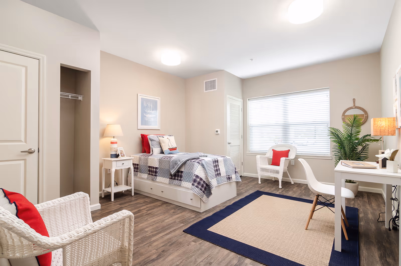A bright and neatly arranged bedroom with a single bed covered in a patchwork quilt and decorative pillows. The room features two white wicker chairs with red cushions, a white desk with a modern chair, a table lamp, a potted plant, and a large window with blinds. The floor is wood with a beige and navy blue area rug.