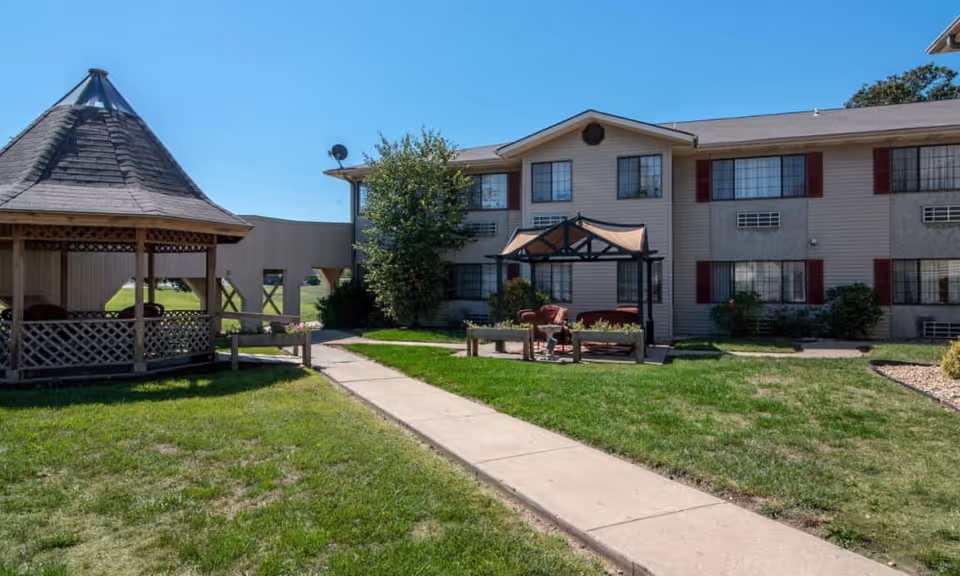 Outdoor view of Harmony Gardens senior living facility showing a two-story building with multiple windows and red shutters. In the foreground, there is a concrete walkway leading to a wooden gazebo on the left and a small covered seating area with chairs and a table on the right, surrounded by green grass and landscaping under a clear blue sky.