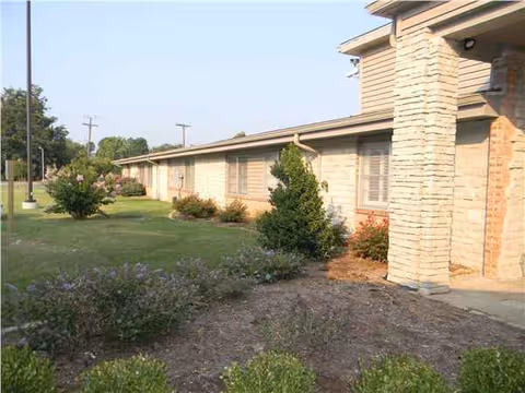 A single-story building with beige stone facade, windows, and landscaped shrubs along the side.
