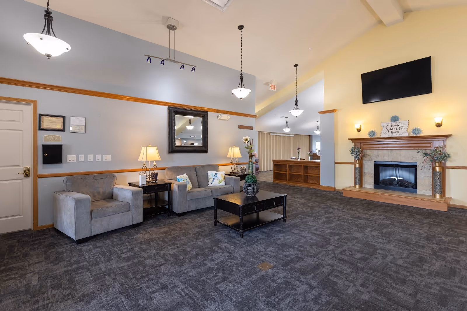 A spacious lobby with sofas and armchairs around a coffee table, a fireplace beneath a wall-mounted TV, and a reception desk in the background.