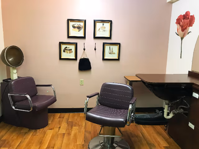 Interior of a salon area with two purple salon chairs, one with a hair dryer hood attached. The room has wooden flooring, a pink wall with four framed pictures, two hooks with a small black bag hanging, and a large red flower decal on the right wall. There is also a black sink with a countertop on the right side.