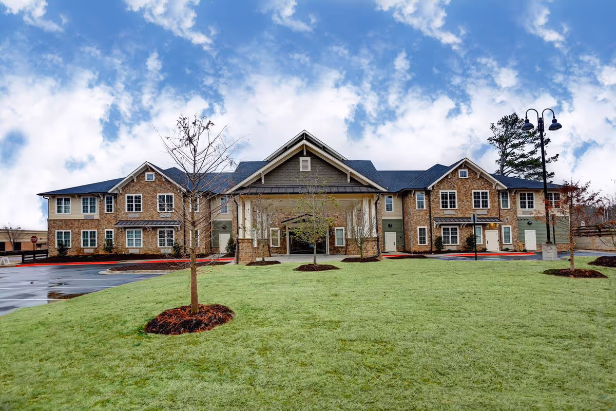 Front exterior view of a two-story senior living facility building with a covered entrance, multiple windows, a green lawn with young trees, and a partly cloudy sky above.