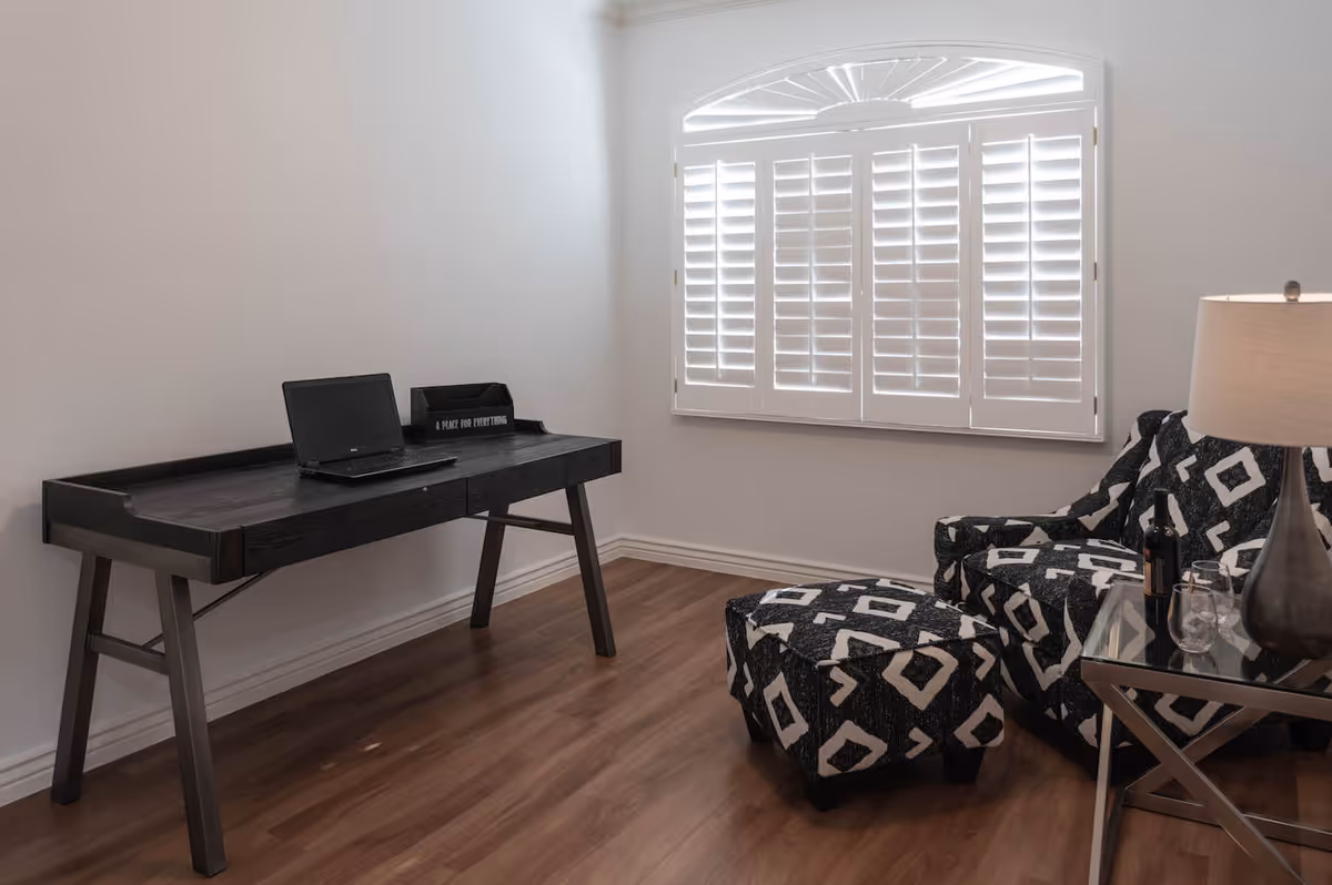 A small room with a wooden desk holding a closed laptop and a black organizer labeled 'A PLACE FOR EVERYTHING'. There is a patterned armchair with a matching ottoman, a glass side table with a lamp, a bottle of wine, and two wine glasses. The room has wooden flooring and a window with white plantation shutters.