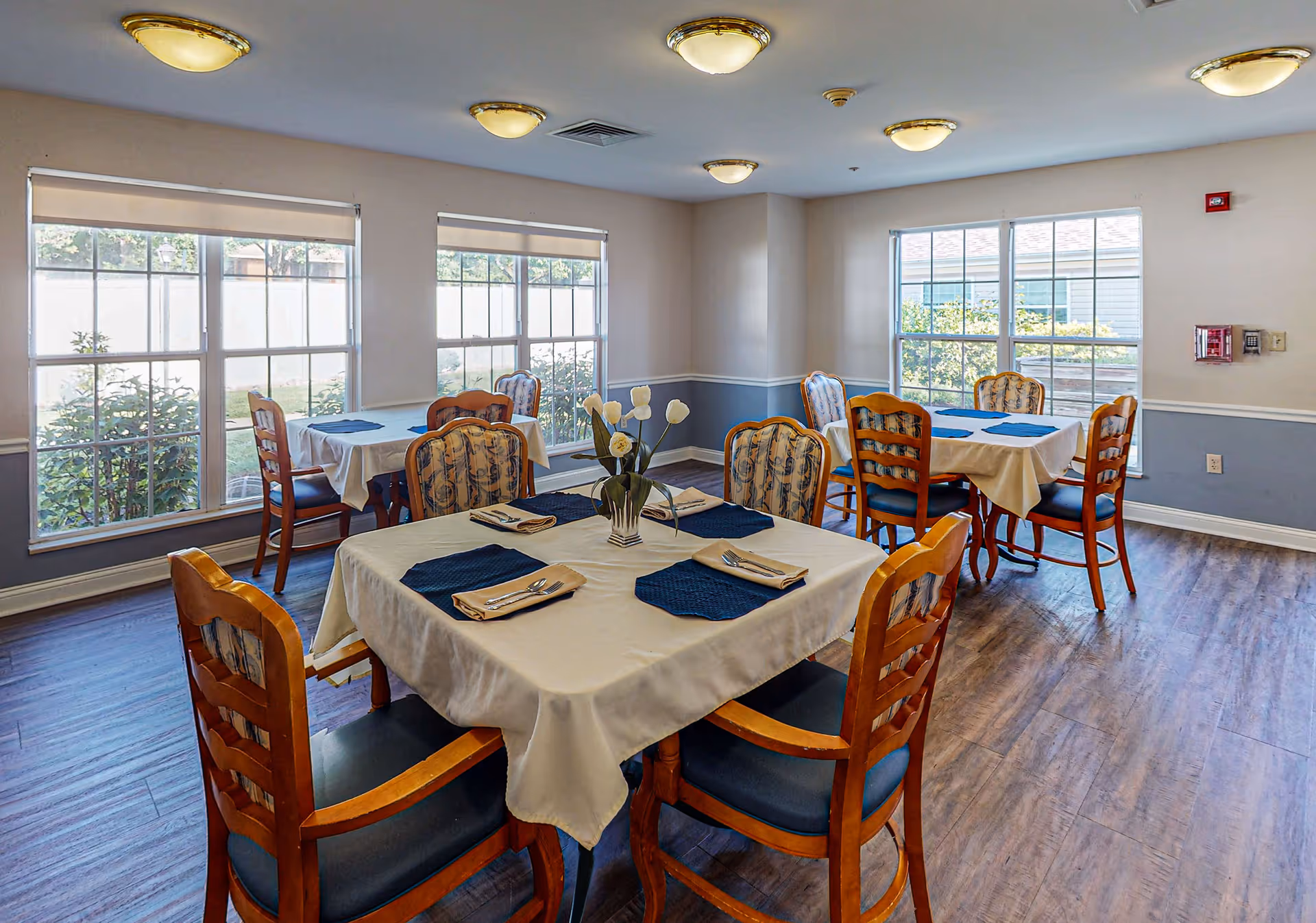 A bright dining room with four tables covered in white tablecloths and blue placemats. Each table is set with napkins and silverware. The room has large windows letting in natural light and wooden chairs with cushioned seats and backs. The floor is wooden, and the walls are painted light blue and white with a chair rail.