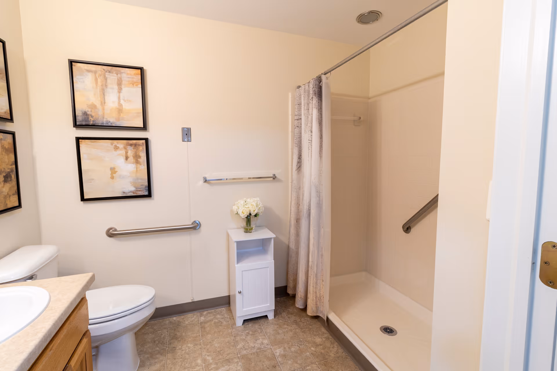 A clean bathroom featuring a toilet, a shower with a curtain, a small white cabinet with a vase of white flowers on top, and two abstract paintings on the wall. The bathroom has beige walls and tiled flooring.