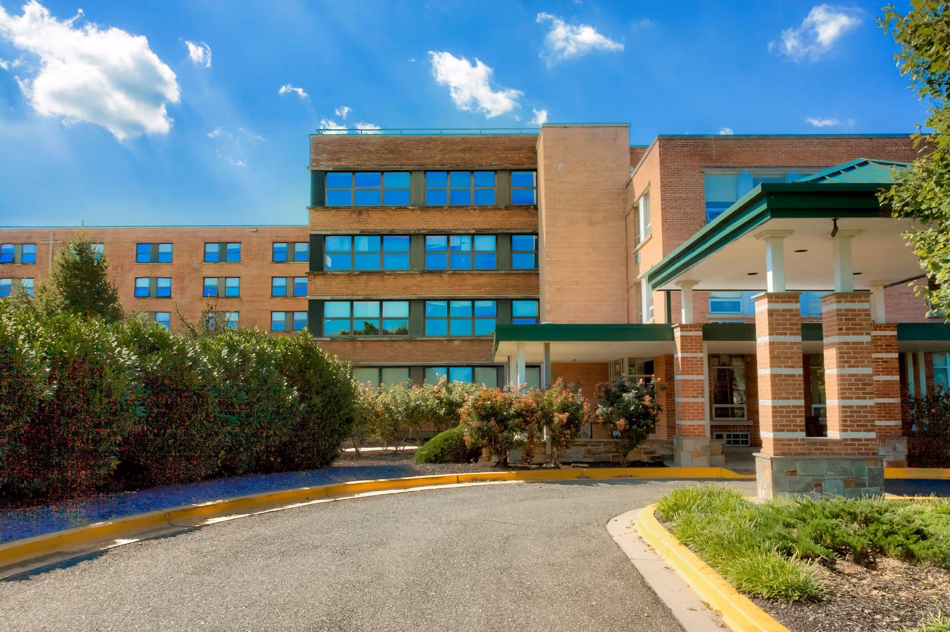 Exterior view of a multi-story brick building with large windows under a blue sky with some clouds. The building has a covered entrance supported by brick columns and is surrounded by green bushes and landscaping.