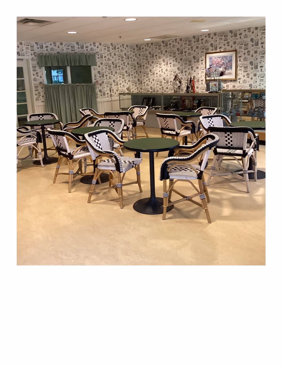 A dining area with multiple small round tables and wicker chairs with black and white woven patterns. The room has light-colored flooring, wallpaper with a vintage print, and a green curtain covering a window. There is a glass display case along the back wall with various decorative items and a framed floral painting above it.