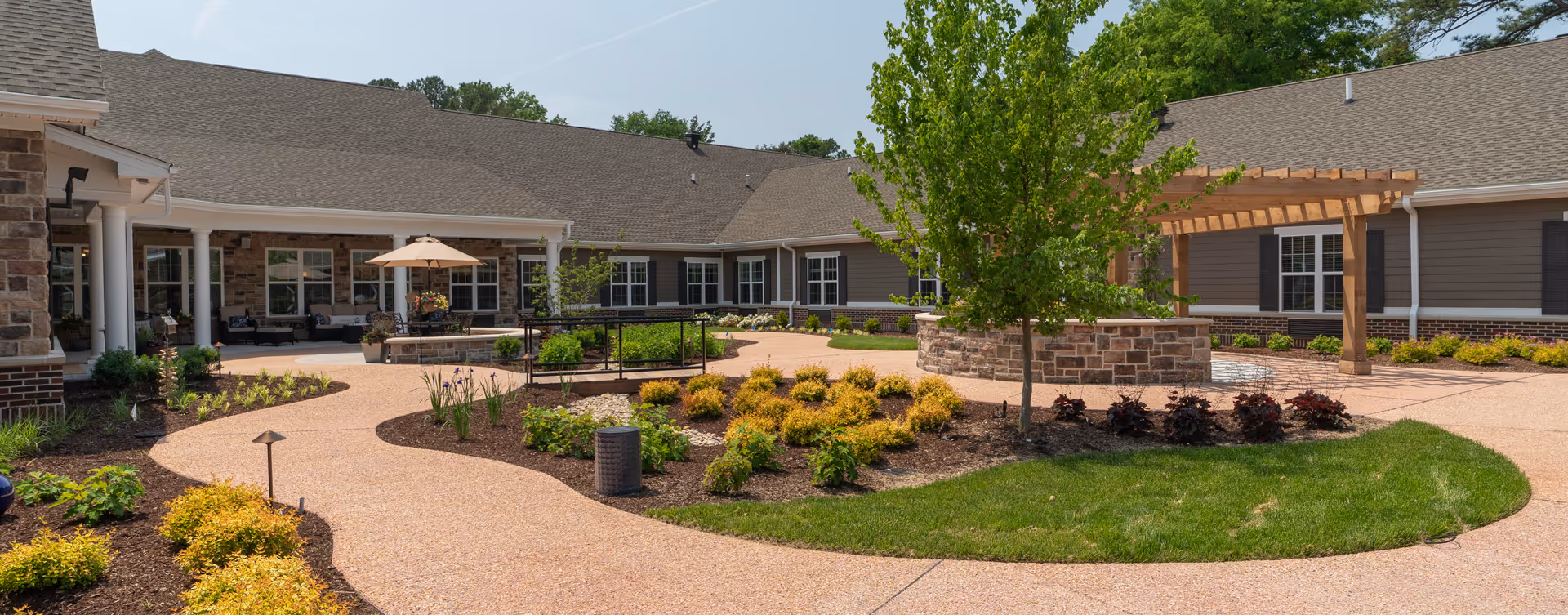 Sunny landscaped courtyard with winding pathways, seating, a pergola and a circular stone planter in front of a single-story senior living building.