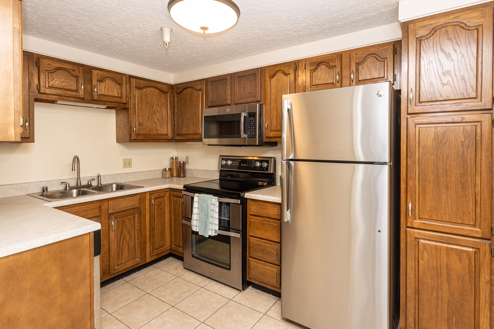 Well-lit kitchen with wooden cabinets, stainless steel refrigerator and stove, a double sink, and tiled floor.