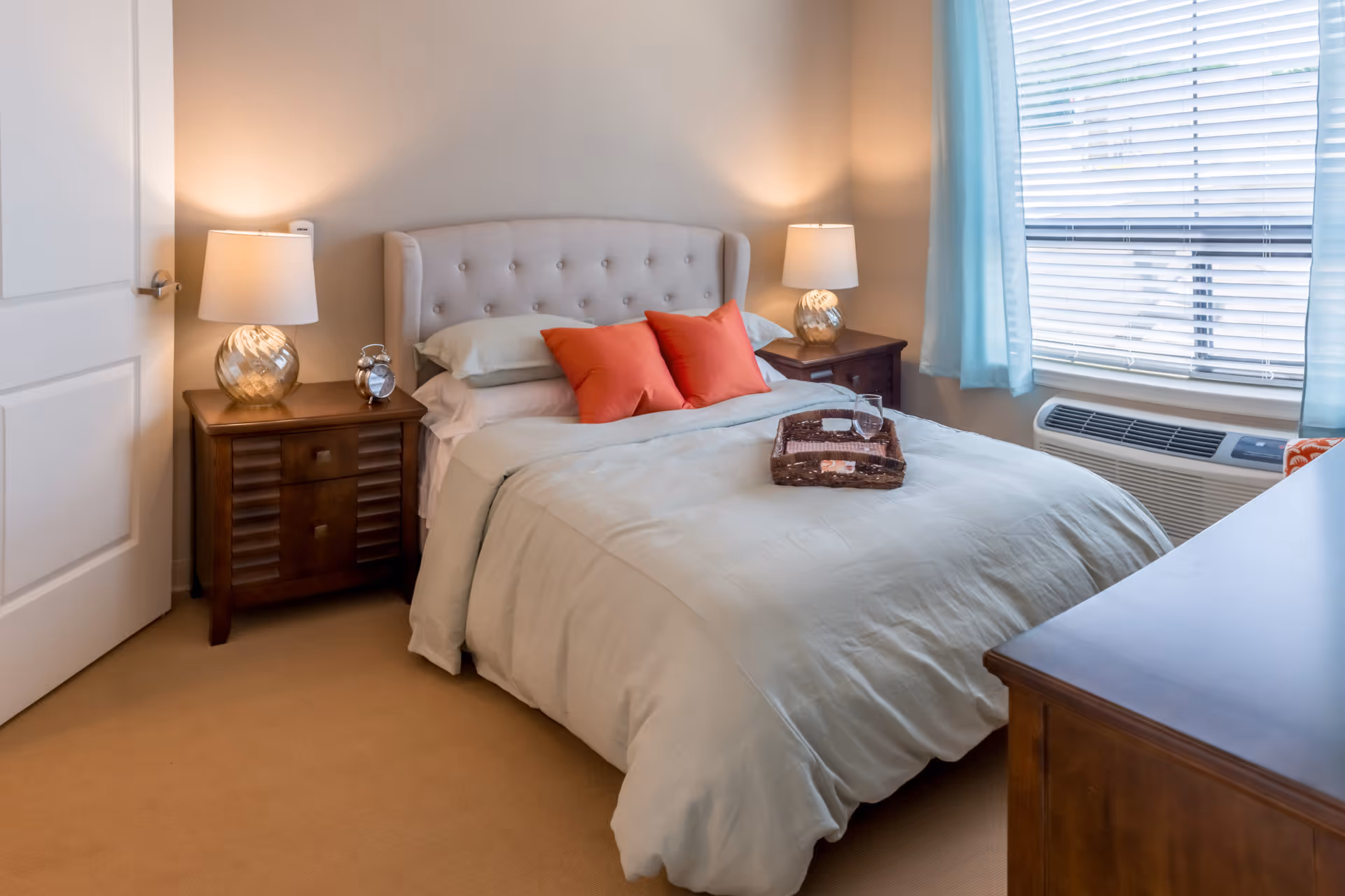 Neatly made bedroom with a tufted headboard, two nightstands with lamps, a window with blinds and light blue curtains, and a dresser.
