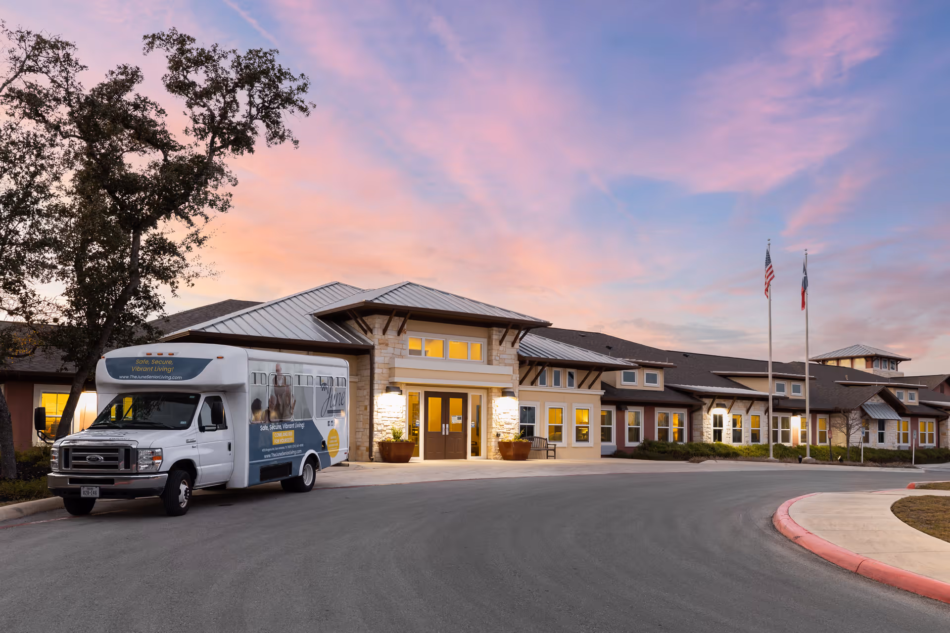 Front exterior of the Waterford at Westover Hills senior living building at sunset with a shuttle van parked by the entrance and flagpoles nearby.