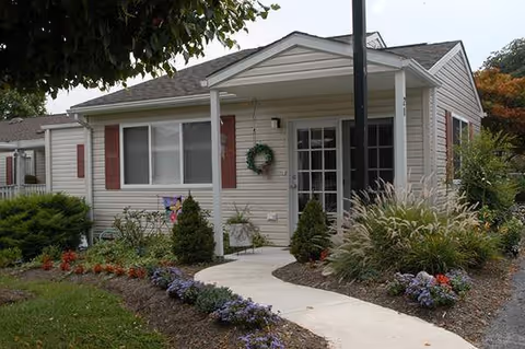 Single-story house with beige siding and red shutters, featuring a small covered porch with a wreath on the door. The front yard is landscaped with various shrubs, flowers, and a curved concrete walkway leading to the entrance.