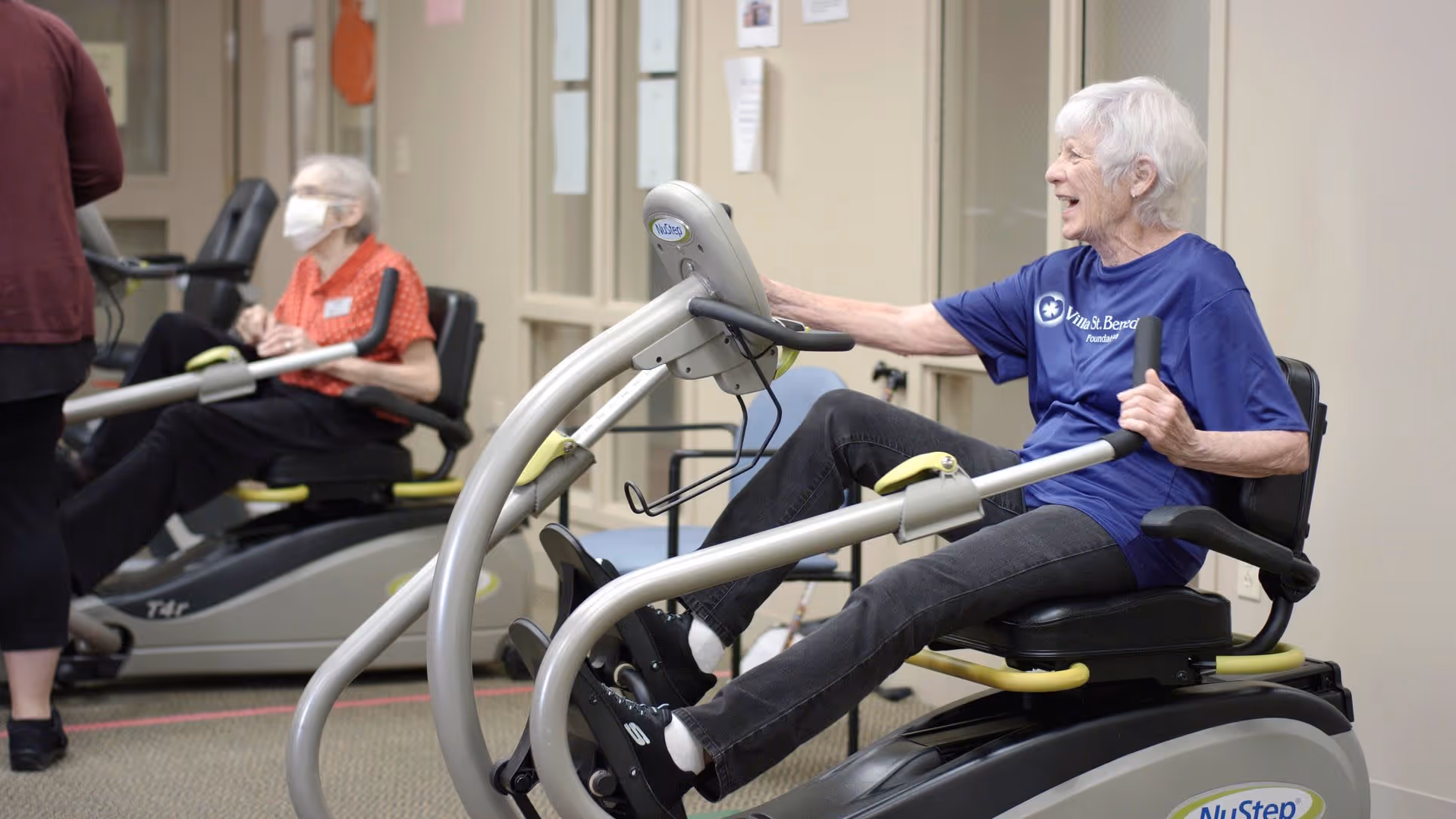 Two elderly women exercising on seated fitness machines in an indoor facility. One woman in a blue Villa St. Benedict t-shirt is smiling while using a NuStep machine, and another woman wearing a face mask is exercising in the background.