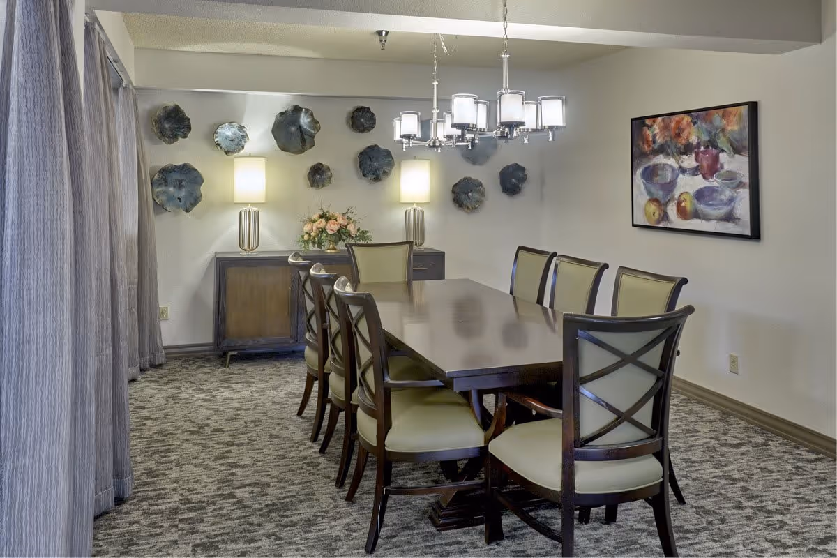 A dining room with a long wooden table surrounded by eight chairs with light-colored cushions. The room features a carpeted floor, two table lamps on a sideboard against the back wall, decorative wall art resembling abstract flowers, and a colorful framed painting on the right wall. A modern chandelier hangs above the table, and sheer curtains cover the window on the left side.