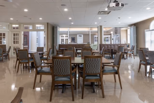 A spacious dining room in Peabody Retirement Community featuring multiple wooden tables and chairs with patterned upholstery. The room has large windows allowing natural light, a polished floor, and a ceiling-mounted projector. The walls are painted in neutral tones with decorative elements and glass-paneled doors.