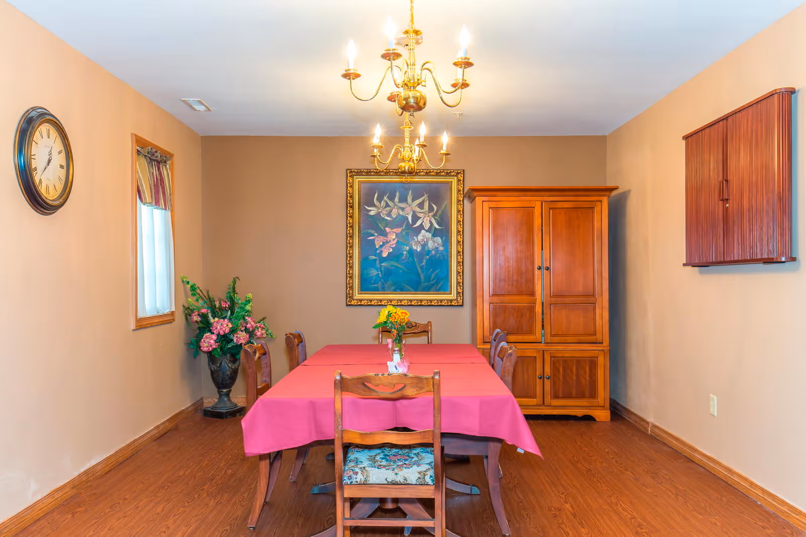 A dining room with a rectangular table covered with a red tablecloth and six wooden chairs. A vase with flowers is placed in the center of the table. The room has wooden flooring, beige walls, a large floral painting in a gold frame on the back wall, a wooden cabinet, a wall-mounted wooden box, a wall clock, a window with a curtain, and a chandelier with lit bulbs hanging from the ceiling.