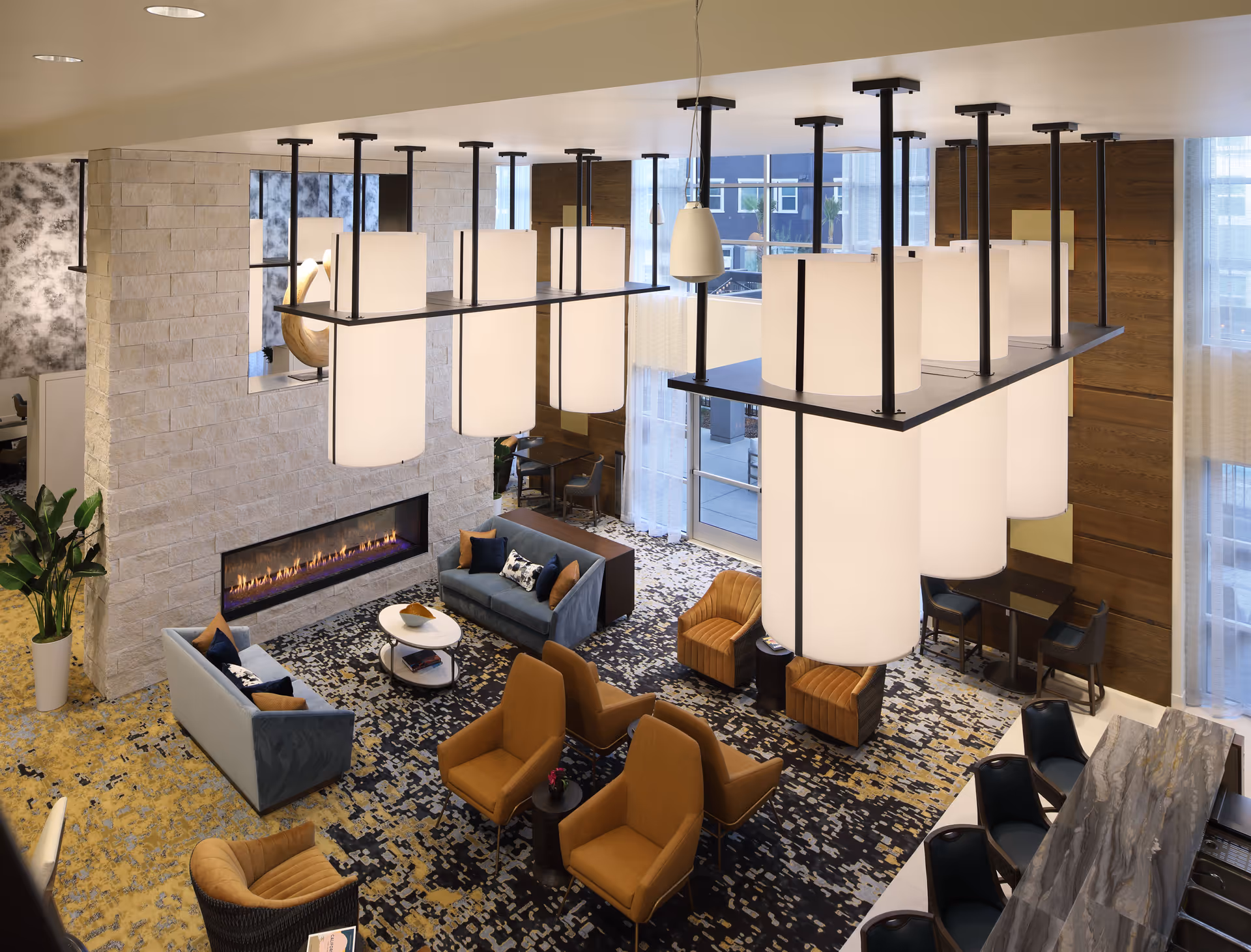 A modern senior living facility common area with a cozy seating arrangement including blue sofas, mustard yellow armchairs, a fireplace set in a stone wall, large hanging cylindrical light fixtures, patterned carpet, and a marble countertop with black chairs.