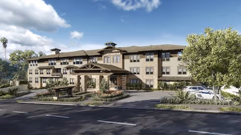 Exterior view of a three-story senior living facility building with a covered entrance, surrounded by landscaping and a parking area with several cars. The sky is partly cloudy.