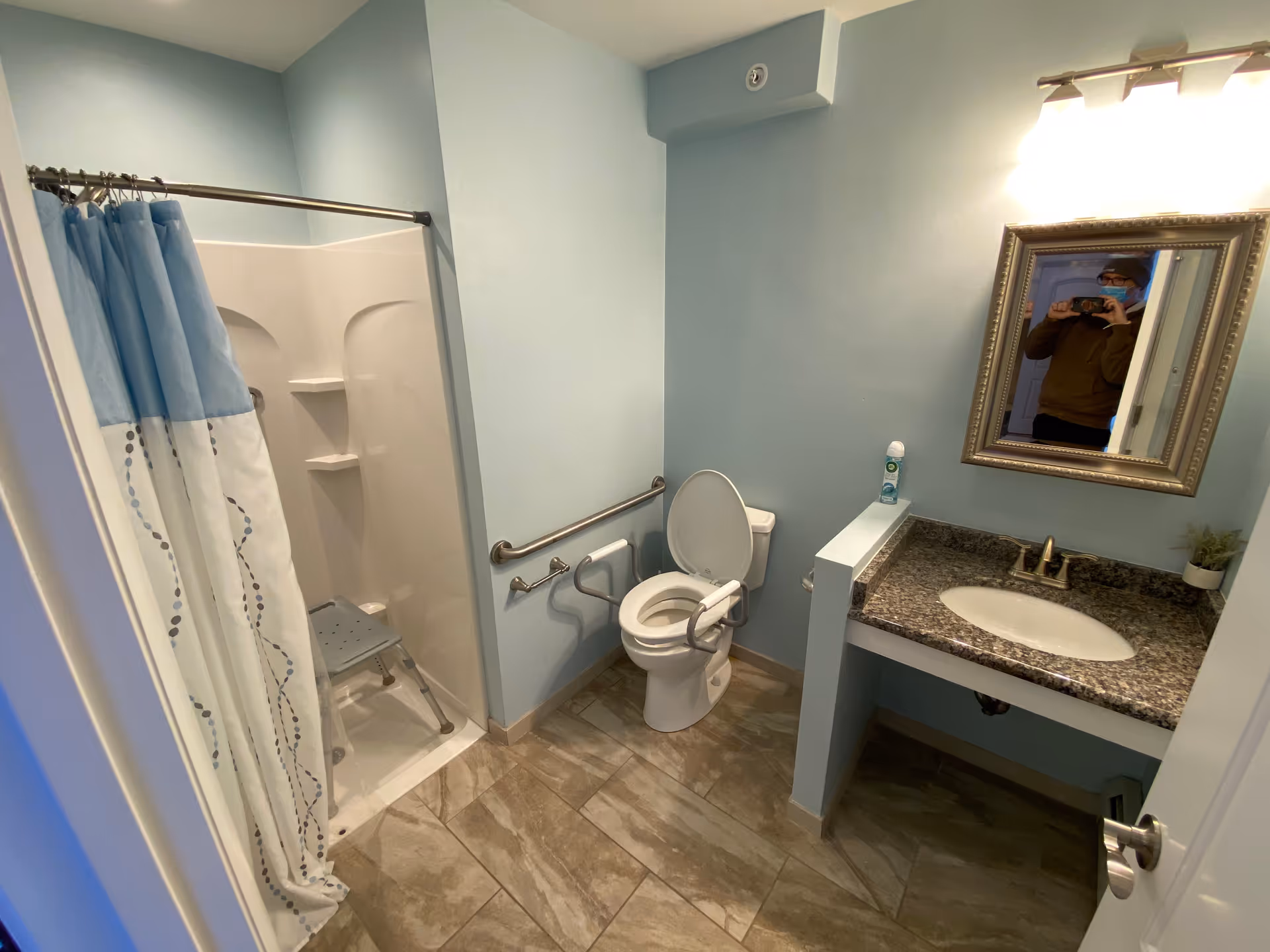 Accessible bathroom with a walk-in shower and shower chair, toilet with grab bars, sink and mirror, and tiled floor.