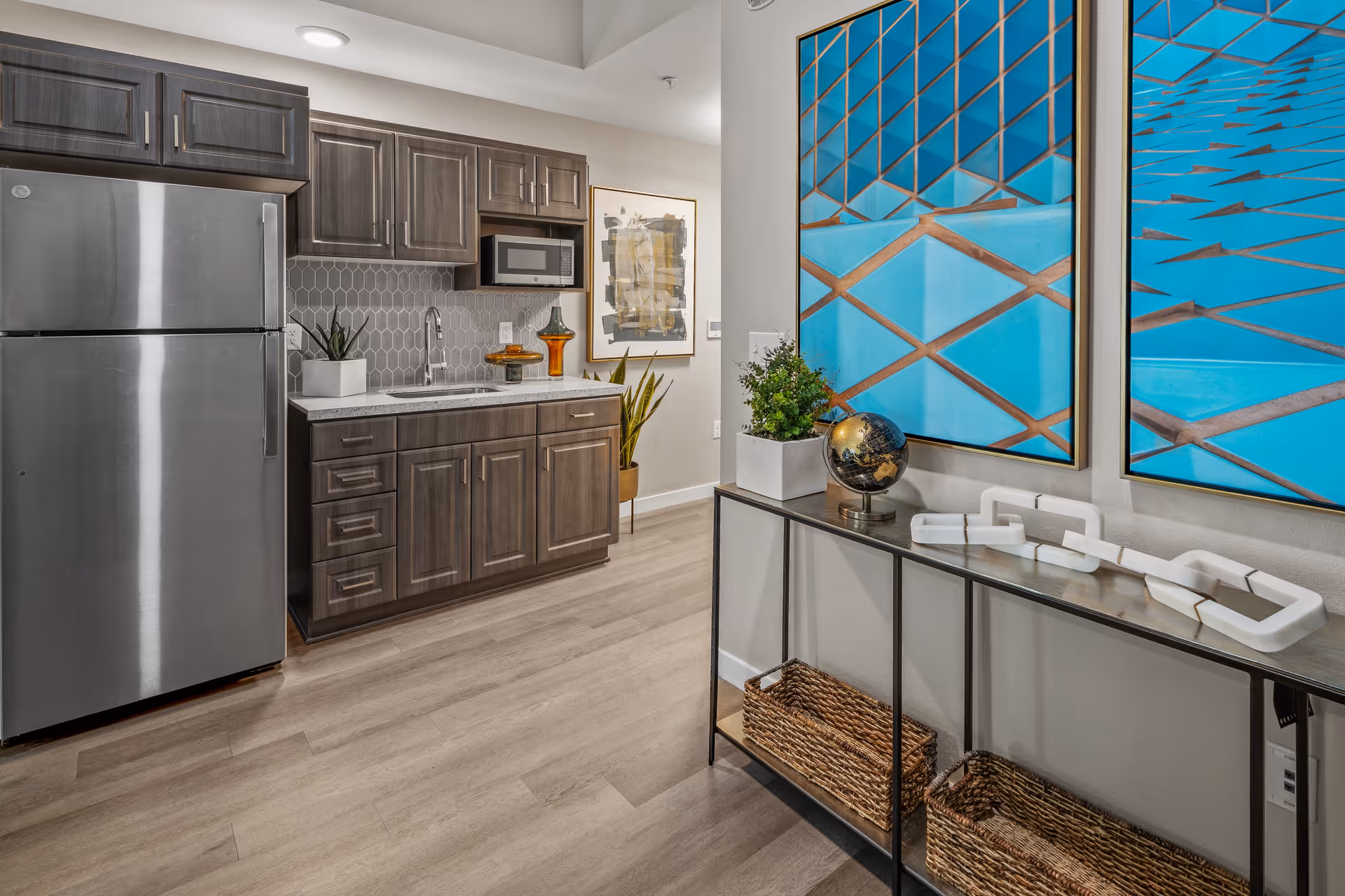 Modern kitchenette with stainless refrigerator, wood cabinets, a sink and microwave, and a console table topped with decorative artwork and plants.