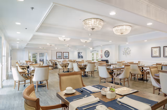 A bright, spacious dining room with neatly set tables, upholstered chairs, and crystal chandeliers.