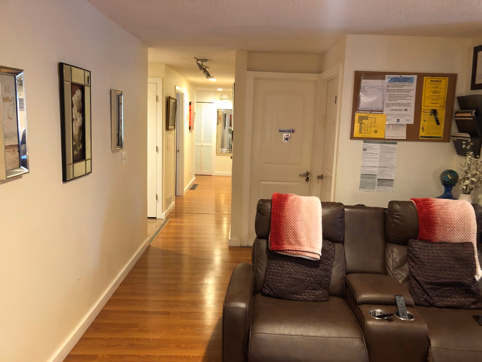 Interior view of a senior living facility hallway with wooden flooring, white walls decorated with framed pictures and mirrors. To the right, there is a brown leather reclining sofa with red and pink blankets draped over the backrests. A bulletin board with various notices and papers is mounted on the wall above the sofa.