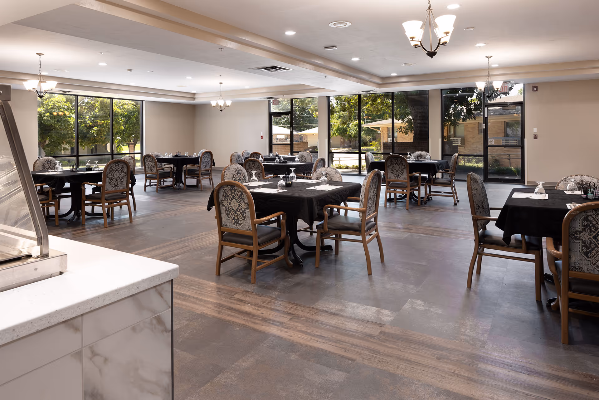 A spacious dining room with multiple tables covered in black tablecloths, each surrounded by wooden chairs with patterned upholstery. Large windows allow natural light to fill the room, and there are chandeliers hanging from the ceiling. The floor has a mix of wood and tile patterns, and there is a counter with a marble-like surface in the foreground.