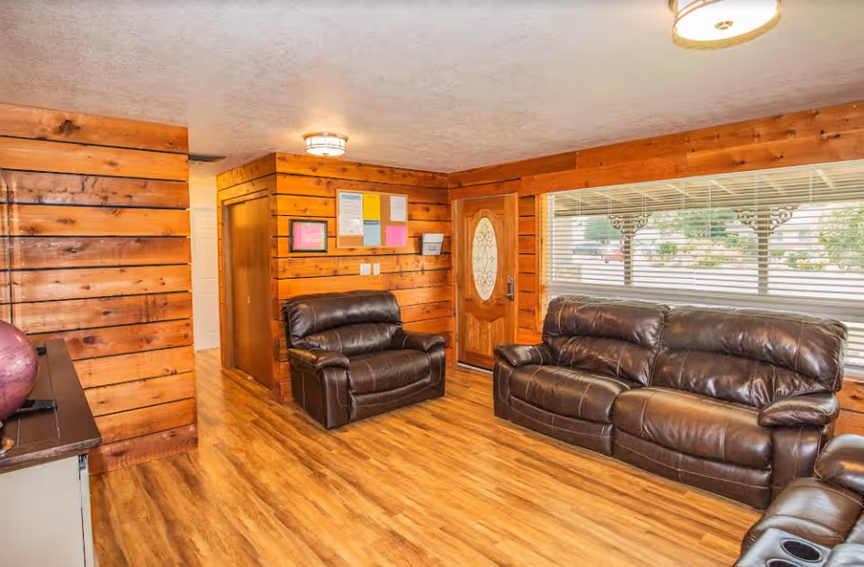 A cozy living room with wooden paneled walls and wood flooring. The room features a brown leather sofa, a matching loveseat, and a recliner. A wooden front door with a decorative glass panel is visible, along with a large window with blinds showing an outdoor view. There are ceiling lights and a bulletin board on the wall near the door.