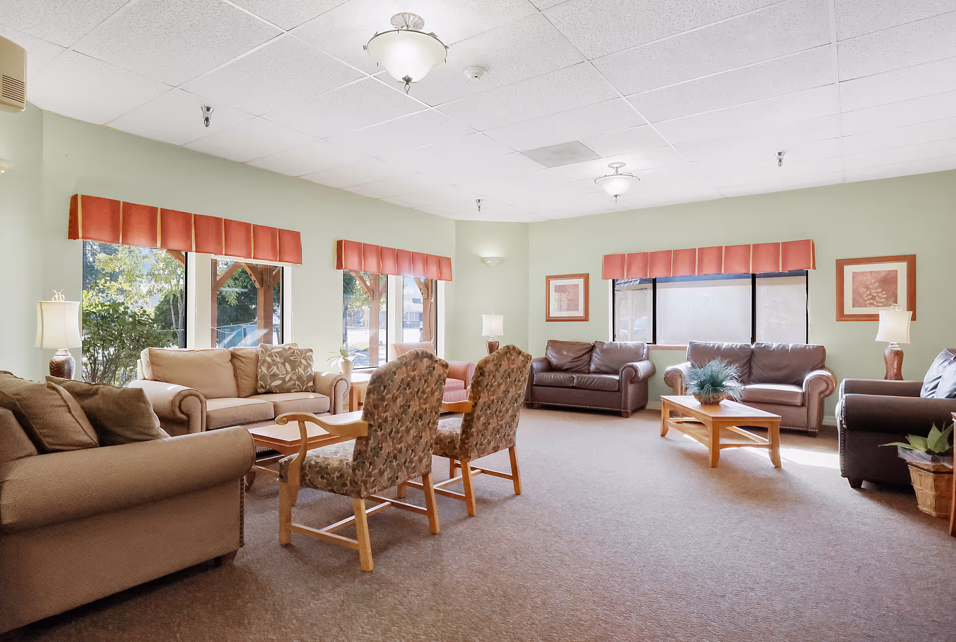 A spacious living room with multiple seating options including beige and brown sofas, two patterned armchairs, and wooden coffee tables. The room has light green walls, large windows with red valances, framed artwork, and several lamps providing soft lighting.