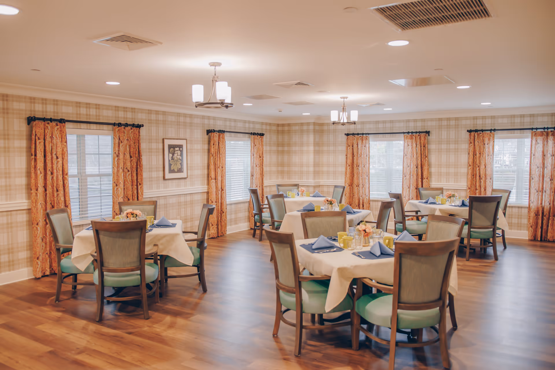 A dining room with several round tables covered with white tablecloths, each set with blue napkins, yellow mugs, and glassware. The room has wooden floors, plaid wallpaper, orange patterned curtains on multiple windows, and ceiling lights.