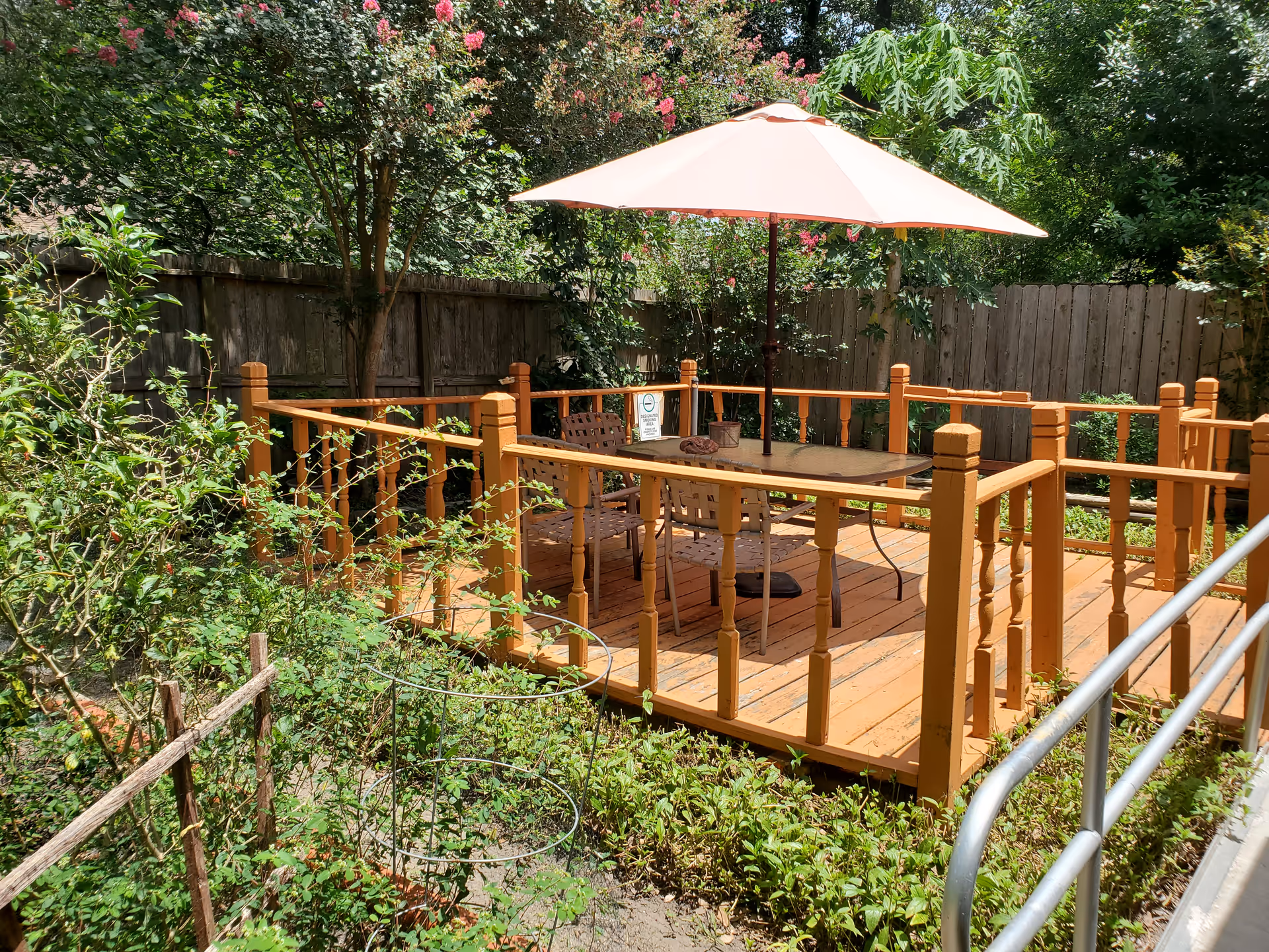 A wooden outdoor deck with a table, chairs and a pink umbrella surrounded by plants and a wooden fence.