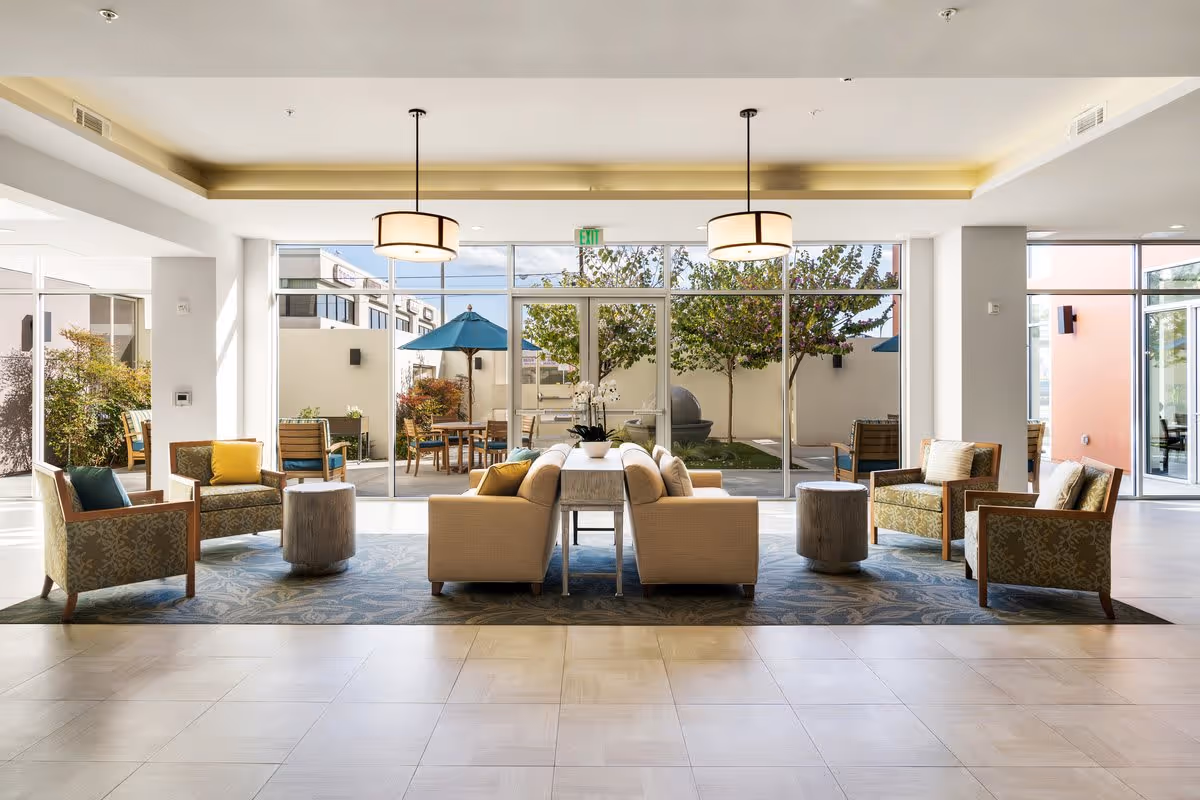 Spacious, sunlit lounge with sofas and armchairs arranged around a rug facing large floor-to-ceiling windows overlooking an outdoor patio.