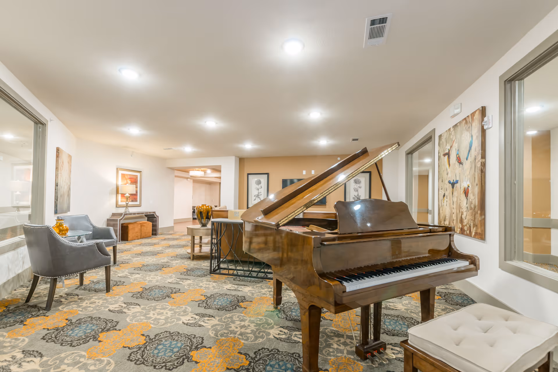 A spacious, well-lit interior room featuring a grand piano with an open lid and a cushioned piano bench. The room has patterned carpet with orange, blue, and gray designs. There are two gray armchairs with a small table and decorative orange vase on the left side. The walls are adorned with framed artwork, including a painting of birds on the right wall. Large windows with gray frames are visible on both sides of the room.