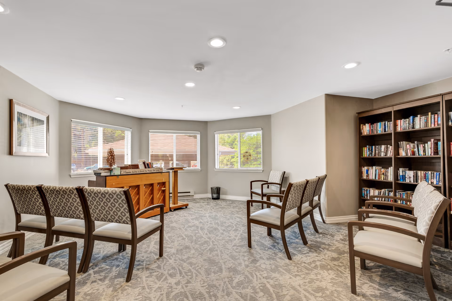 A bright and spacious room with several chairs arranged in a semi-circle facing a wooden piano and a lectern. The room has large windows with blinds letting in natural light, a bookshelf filled with books on the right side, and a framed picture on the left wall. The carpet has a subtle pattern and the walls are painted light beige.