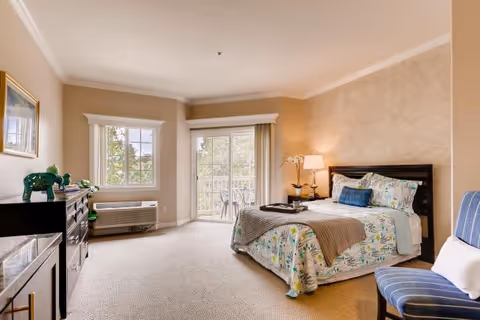 A bright and spacious bedroom with a large bed featuring floral bedding and a blue pillow. There is a nightstand with a lamp and a decorative orchid next to the bed. The room has beige walls and carpeted flooring. A dresser with decorative items is on the left side, and a blue striped chair with a white pillow is on the right. Two windows and a sliding glass door leading to a balcony allow natural light to fill the room.