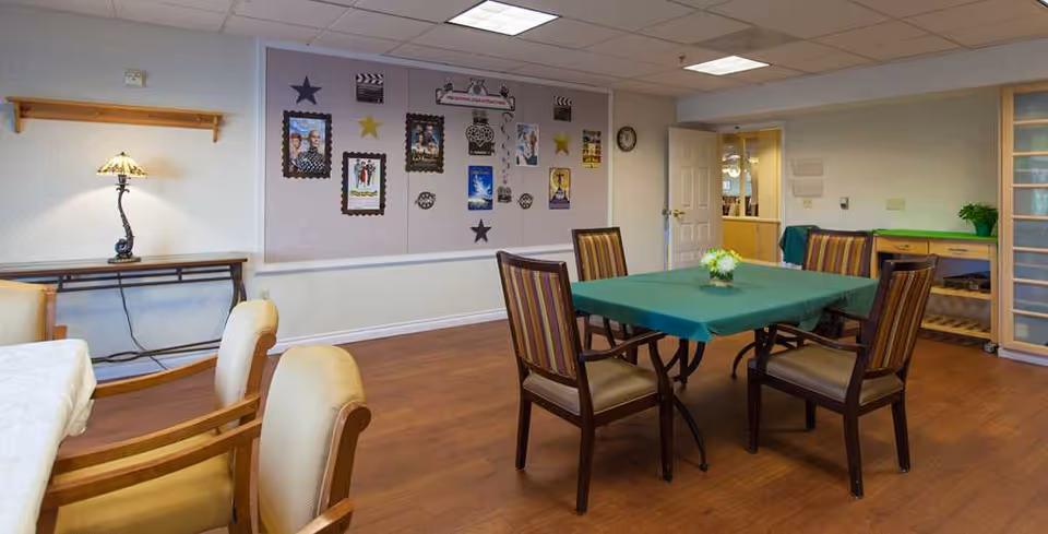 A well-lit room with wooden flooring featuring two tables covered with tablecloths, surrounded by chairs. One table has a green tablecloth with a small flower arrangement in the center. The walls have decorative framed pictures and stars, and there is a lamp on a side table. An open door leads to another room with visible shelving and a clock on the wall.