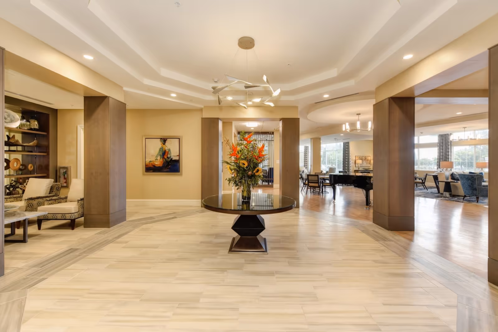 A spacious and well-lit interior common area of a senior living facility with a round table in the center holding a large floral arrangement. The room features beige walls, tiled flooring, modern ceiling lights, and wooden pillars. To the right, there is a lounge area with chairs, tables, and a grand piano near large windows allowing natural light to fill the space. On the left, there are comfortable chairs and decorative shelves with art pieces.