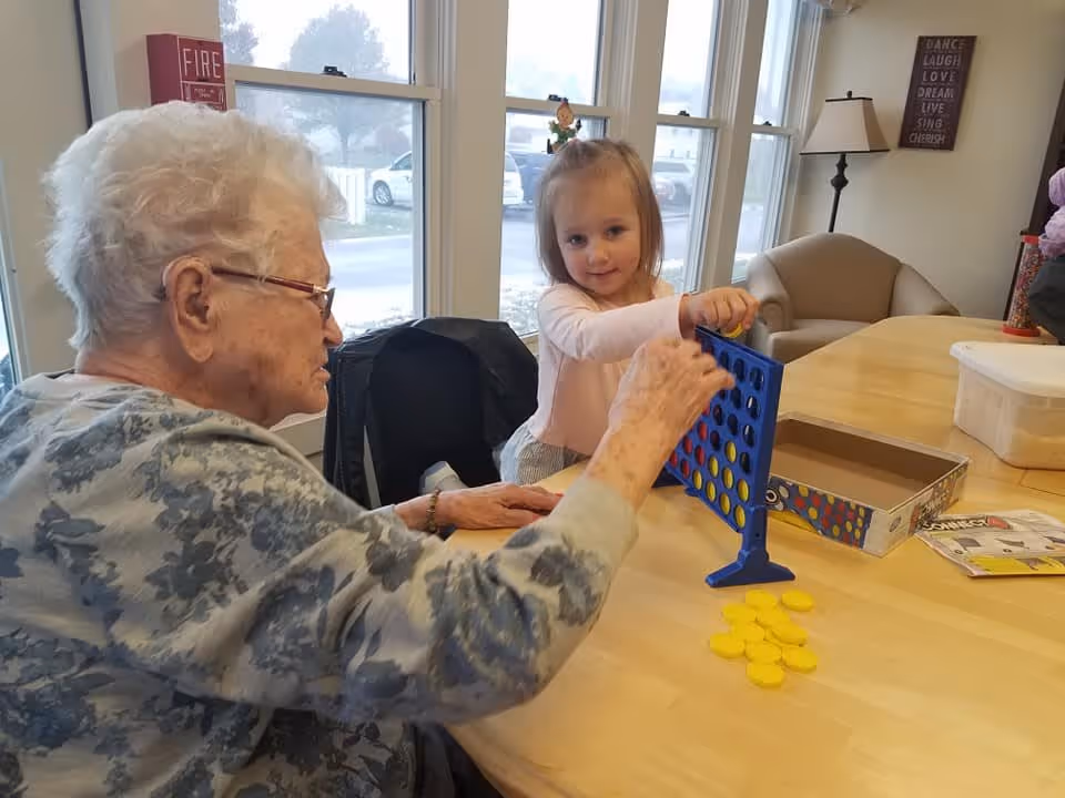 An elderly woman and a young girl playing a game of Connect Four at a wooden table in a bright room with large windows. The elderly woman is wearing glasses and a floral patterned sweater, while the girl is smiling and placing a yellow game piece into the blue game grid. In the background, there is a beige armchair, a floor lamp, and a wall decoration with inspirational words.