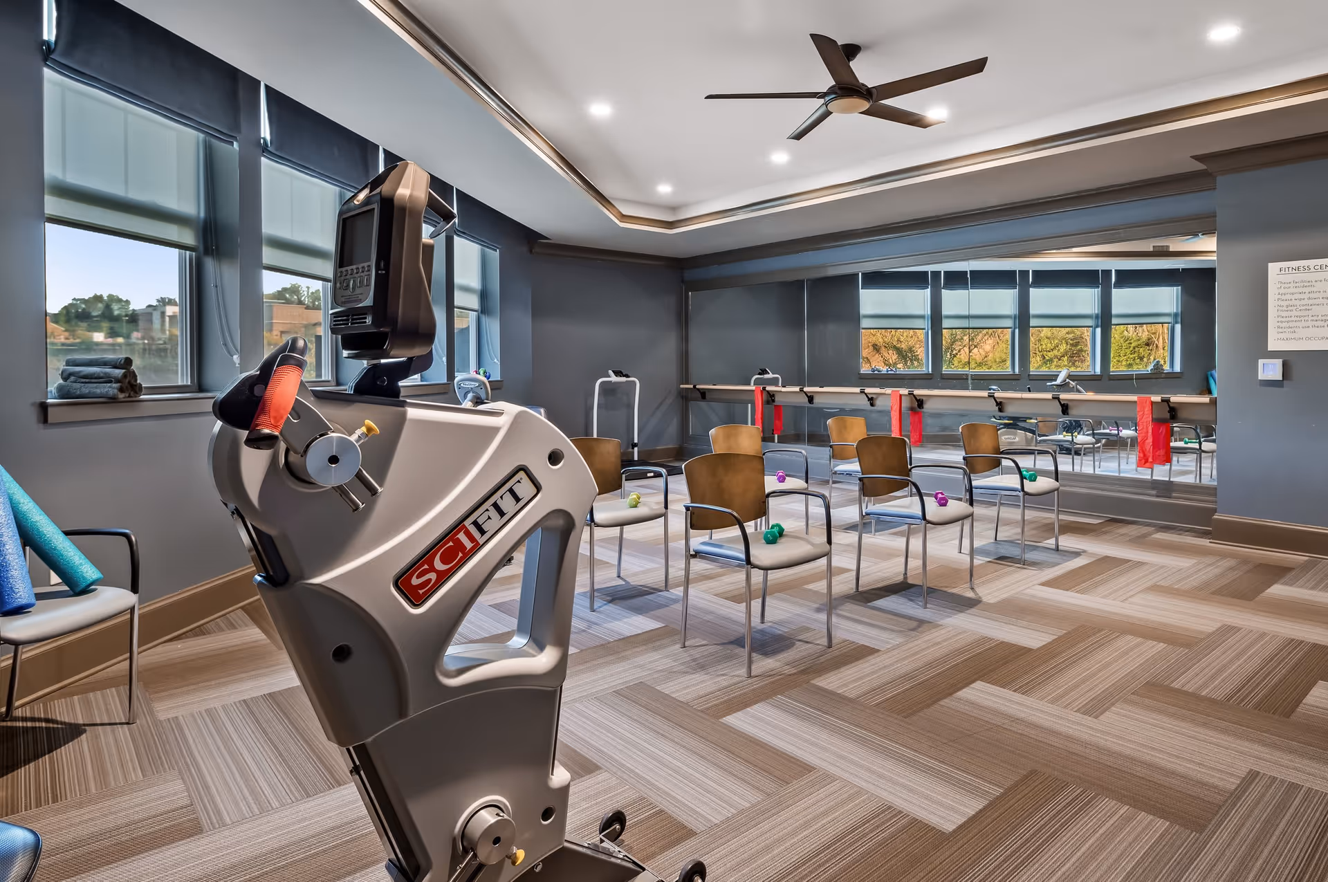 A fitness room with a SciFit exercise machine in the foreground, several chairs arranged in rows each with a small dumbbell on the seat, a large mirror on the wall, windows with shades partially down, and a ceiling fan above.